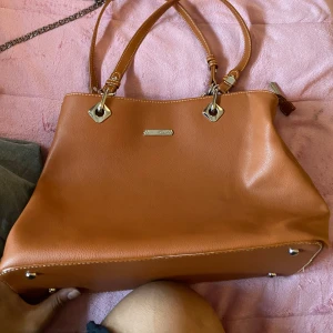 Brun axelväska i skinn - Säljer en snygg brun axelväska i skinn. Väskan har två handtag och en avtagbar axelrem. Den har en dragkedja på baksidan och flera fack inuti för bra förvaring. Perfekt för både vardag och fest!