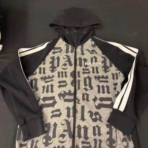 Palm Angels hoodie med tryck och huva - Säljer en snygg hoodie från Palm Angels med svart och grå färg, stora bokstavstryck över hela framsidan och vita ränder på ärmarna. Tröjan har huva, dragkedja och långa ärmar. Perfekt för dig som gillar streetwear och vill sticka ut.
