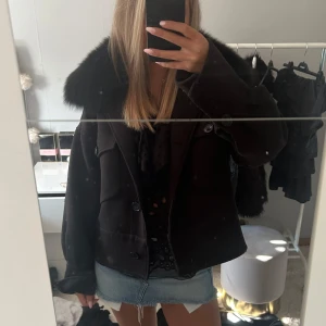 Meotine jacka - Snygg svart jacka med stor, fluffig fuskpäls-krage och knappar framtill. Jackan har en boxig passform och långa ärmar, perfekt för kyligare dagar. Stilsäker och trendig att bära till både jeans och kjo