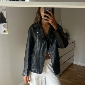 Svart skinnjacka med dragkedjor - Säljer en svart skinnjacka med cool bikerstil. Jackan har silvriga dragkedjor, snedställd front och klassisk krage med tryckknappar. Långärmad modell med fickor och detaljer vid ärmslut. Perfekt för dig som vill ha en edgy look.