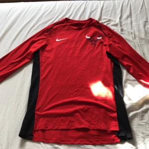 Röd långärmad Nike Bulls tröja - Säljer en autentisk röd långärmad tröja från Nike med Chicago Bulls-logga på bröstet och svart panel längs sidorna. Tröjan har rund hals och är gjord i mjukt polyester-material, perfekt för basket eller chill. NBA-logga på ryggen för extra street cred.