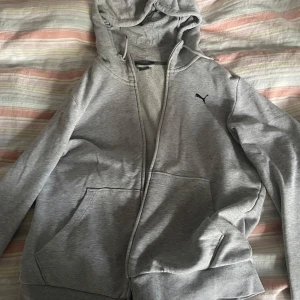 Grå hoodie från Puma med dragkedja - Säljer en grå hoodie från Puma i storlek M. Tröjan har huva, långa ärmar, två stora fickor framtill och en svart Puma-logga på bröstet. Materialet är mjukt och känns som bomull och polyester. Perfekt för chill dagar eller när du vill ha en enkel streetstyle.