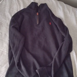 Svart half zip tröja från Polo Ralph Lauren - Svart långärmad tröja från Polo Ralph Lauren med röd broderad logga på bröstet. Tröjan har en half zip-dragkedja vid halsen och är tillverkad i mjukt bomullsmaterial. Perfekt för lager-på-lager och chill stil.
