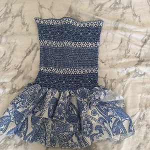 Blåvit kortklänning med volang - Supersöt blåvit kortklänning med smockad överdel och bred volang nedtill. Klänningen har ett detaljerat blommigt mönster och är axelbandslös, perfekt för varma dagar. Materialet känns lätt och luftigt, vilket gör den skön att bära under sommaren. Köpta utomlands❣️❣️