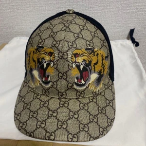 Gucci keps tiger beige - Snygg keps från Gucci med klassiskt GG-mönster i beige och brunt, prydd med två stora tigerhuvuden framtill. Baksidan är i svart mesh och justerbar rem med gröna och röda detaljer. Perfekt statement-piece för dig som vill sticka ut.