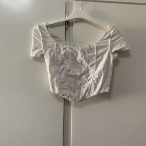 Vit croppad korsett-topp - Säljer en vit croppad korsett-topp med korta ärmar och markerade sömmar framtill. Toppen har en snygg, figurnära passform och en lätt böjd nederkant som ger en cool siluett. Perfekt att styla med jeans eller kjol för en trendig look.