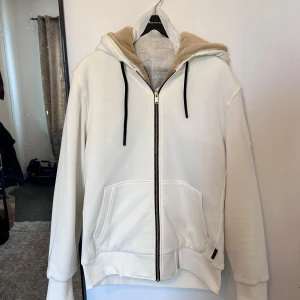 Vit hoodie från Moose Knuckles med päls - Säljer en vit hoodie från Moose Knuckles med guldfärgad logga på ärmen och svart dragkedja. Insidan av huvan är fodrad med mjuk beige päls. Super fin men säljs nu då den enbart tagits på för provning och aldrig kommit till användning.