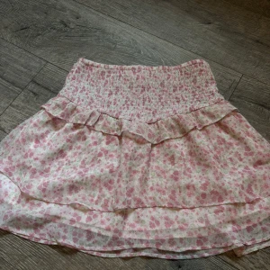 Blommig volangkjol från Gina Tricot - Supersöt kort kjol från Gina Tricot med blommigt mönster i rosa, vitt och lite grönt. Kjolen har smockad midja och volangdetalj upptill, vilket ger en flowy och lätt känsla. Perfekt för dig som gillar romantisk och feminin stil. Aldrig andvönd. 