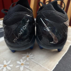 Nike Phantom GT2 Elite fotbollsskor svart/blå - Snygga Nike Phantom GT2 Elite fotbollsskor i svart med blå detaljer och silverfärgad swoosh. Ovandelen är i syntetmaterial med textur för bättre bollkontroll. Skorna har snörning och blå dobbar för optimalt grepp på planen. Perfekta för dig som vill sticka ut på matchen. Obs ( de är använda i 1 år och köptes för 2800 och kommer med dens påse!!