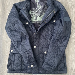 Mörkblå quiltad jacka från Barbour - Snygg mörkblå quiltad jacka från Barbour. Jackan har flera fickor framtill och coolt tryck på insidan. Perfekt för dig som gillar stilren och tidlös design. Den perfekta vinter jackan!