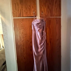Ljuslila långklänning med drapering - Säljer en elegant ljuslila långklänning med smala axelband och snygg drapering över bröstet och längs sidan. Klänningen har en glansig finish och faller mjukt ner mot golvet, perfekt för dig som vill sticka ut med en sofistikerad look.