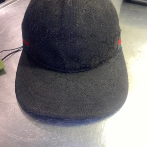 Gucci Keps - Svart keps från Gucci med klassiskt GG-monogram över hela kepsen. Sidorna har grönt och rött band, typiskt för Gucci. Justerbar rem baktill i svart skinn. Snygg och stilren accessoar för dig som vill sticka ut. Inga skavanker.