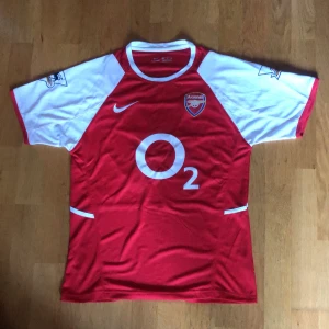 Arsenal Henry 14 Nike fotbollströja M - Säljer en klassisk Arsenal fotbollströja med Henry 14 på ryggen. Tröjan är röd med vita korta ärmar, har Arsenal-emblem, O2 sponsortryck och Barclays Premier League patch. Tillverkad i lätt och ventilerande polyester med Dri-FIT teknologi.