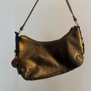 Guldig axelväska från Don Donna - Snygg axelväska i metallic guld från Don Donna. Väskan har en mjuk, rundad form och är tillverkad i skinnimitation🤎