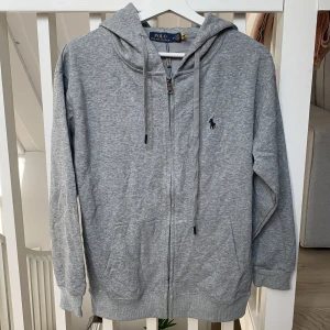 Grå hoodie från Polo Ralph Lauren - Snygg grå hoodie från Polo Ralph Lauren med dragkedja och huva. Tröjan har långa ärmar, snörning vid huvan och klassisk Polo-logga broderad på bröstet. Tillverkad i mjuk bomull för skön komfort. Perfekt för chill dagar eller när du vill ha en clean look.