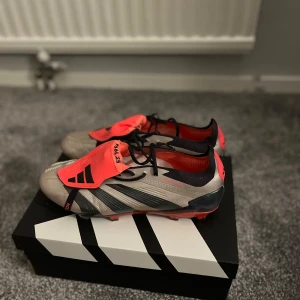 Adidas Predator fotbollsskor silver/röd. Elite - Snygga Adidas Predator fotbollsskor i silver med svarta detaljer och röda inslag. Skorna har snörning, svart Adidas-logga på den röda plösen och en röd yttersula med gräs dobbar.