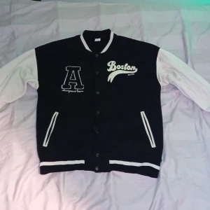 Svart och vit bomberjacka Boston - Collegeinspirerad svart och vit bomberjacka med tryck 'Boston' och stort A på bröstet. Jackan har vita ärmar, svarta muddar med vita ränder och knäppning framtill. Två fickor med vita detaljer. Perfekt för en sportig och avslappnad stil.