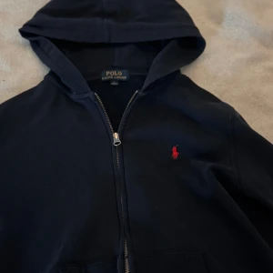 Mörkblå hoodie från Polo Ralph Lauren - Snygg mörkblå hoodie från Polo Ralph Lauren med dragkedja och huva. Tröjan har det klassiska röda Polo-logot broderat på bröstet och är tillverkad i mjuk bomull. Perfekt för chill dagar och enkel att matcha med jeans eller joggers. Fint skick men vissa slitningar 💕