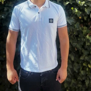 Vit Stone Island piké med svart logga - Vit pikétröja från Stone Island med svart logga på bröstet. Klassisk krage med svarta ränder och knappslå framtill. Kortärmad och tillverkad i mjuk bomull som är skön mot huden. Perfekt för dig som gillar clean och stilren look.                                                                                                                                                                               Nypris: 1999kr GORILLAPRIZE:820kr            Skick:8/10 mycket bra         KVITTO MEDKOMMER     