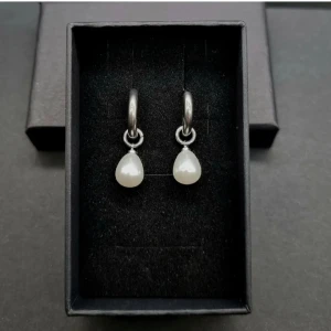 Två par silverörhängen med pärldetaljer - Säljer två par snygga örhängen i silverfärg. Ena paret har droppformade vita pärlor som hänger från runda hoops. Det andra paret har små vita pärlor och coola konformade hängen, med glittriga detaljer på öronkroken. Perfekta accessoarer för att lyfta din stil.