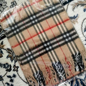 Fleece Halsduk I Klassisk Mönster  - Snygg beige halsduk med klassiskt rutigt mönster i svart, vitt och rött. Mjukt material och långa fransar i ändarna som ger en cool touch. Perfekt att slänga över axlarna för extra stilpoäng när det är kallt ute.