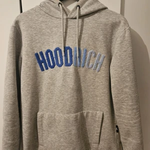 Grå hoodie från Hoodrich med blå text - Snygg grå hoodie från Hoodrich i storlek small. Tröjan har broderad blå och ljusblå text på bröstet, huva med snörning och en stor magficka. Perfekt för dig som gillar streetwear och vill ha en chill look.