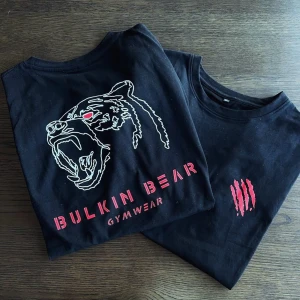 Svart BulkIn Bear t-shirt med tryck - Svart t-shirt från BulkIn Bear med coolt tryck av en björn med röda detaljer och texten 'GYMWEAR' på framsidan. På den andra t-shirten syns röda klomärken. Perfekt för gymmet och streetwear. Rund hals och korta ärmar.