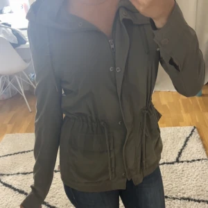 Olivgrön parkajacka från ONLY - Snygg olivgrön parkajacka från ONLY med dragsko i midjan, stora fickor och huva. Jackan har dragkedja och knappar framtill samt långa ärmar. Perfekt för vår och höst när du vill ha en chill och stilren look.