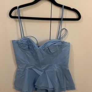 Blå korsettopp med volang från Zara - Supersöt ljusblå korsettopp från Zara med tunna axelband, volang nertill och rynkade kupor. Toppen har smock i ryggen och knytband i sidorna för extra detalj. Perfekt att styla med jeans eller kjol för en trendig look.