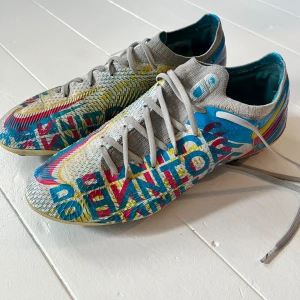 Nike färgglada fotbollsskor med snörning - Säljer ett par Nike fotbollsskor med färgstarkt mönster i blått, rött, gult och grått. Skorna har snörning, stickad ovandel och plös, samt dobbar för bra grepp på planen. Perfekta för dig som vill sticka ut på fotbollsplanen. Nypris- 2799