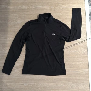 Svart half zip tröja från J.Lindeberg - Svart långärmad tröja från J.Lindeberg med stilren vit logga på bröstet. Tröjan har en hög krage och half zip-dragkedja framtill. Materialet är mjukt och stretchigt, perfekt för träning eller chill. Snygg passform som sitter bra utan att vara för tight. Storlek X-xs