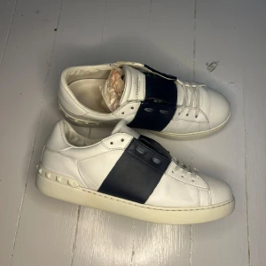 Valentino Garavani Open sneakers vit/navy - Snygga Valentino Garavani Open sneakers i vitt skinn med bred navy läderdetalj över snörningen och ikoniska nitar på hälen. Klassisk rund tå och platt sula. Perfekta för dig som gillar exklusiva och stilrena sneakers med en twist. Skorna är i storlek 41 och i bra skick! För fler frågor kom privat!