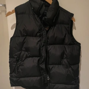 Svart dunväst från Gant Woman - Säljer en svart dunväst från Gant Woman med hög krage och dragkedja med G-detalj. Västen är quiltad och har två fickor med dragkedja. Yttermaterialet är nylon och den är fylld med dun, perfekt för lager-på-lager under kyliga dagar.