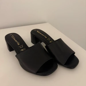 Svarta klackade sandaler från Tamaris - Stilrena svarta sandaler från Tamaris med bred rem över foten och öppen tå. Skorna har en stabil och låg klack så väldigt bekväma att gå i. Har tyvärr aldrig kommit till användning. Skorna är i storlek 38 men skulle säga att det även passar för dig med storlek 37. 