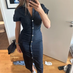 Mörkblå jeansklänning från Zara - Supersnygg mörkblå jeansklänning från Zara med knappar hela vägen framtill, v-ringning och korta ärmar. Klänningen har figurnära passform, krage och en liten slits nertill. Perfekt för dig som gillar en stilren och trendig look.