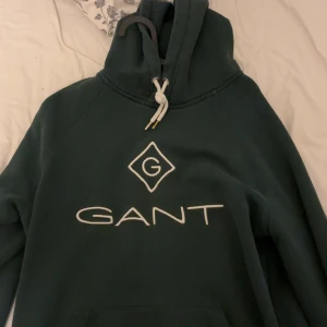 Mörkgrön hoodie från GANT - Snygg mörkgrön hoodie från GANT med stor vit logga framtill. Tröjan har huva med vit snörning och långa ärmar. Perfekt för en chill och avslappnad stil. Klassisk passform och mjukt material.