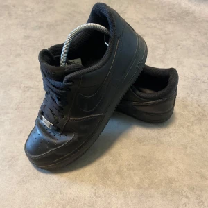 Svarta Nike Air Force 1 sneakers - Klassiska helsvarta Nike Air Force 1 sneakers i skinn med lågt skaft och rund tå. Skorna har perforerad tåbox, svart sula och snörning. Perfekta för dig som gillar en clean och tidlös streetstil. De är i fint skick förutom att sulan är lite trasig och sliten men inget som märks eller syns vid användning | kolla gärna in profilen för liknande produkter och skriv vid minsta lilla fråga 😁😁