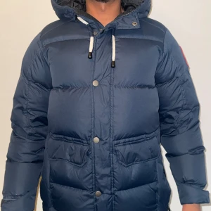 Blå dunjacka med huva från Tuxer XXS - Säljer en mörkblå dunjacka från Tuxer i storlek XXS. Jackan har huva med snörning, stora fickor framtill och stängs med både dragkedja och knappar. Yttermaterialet är polyester och fyllningen består av 70% dun och 30% fjäder. Perfekt för kalla dagar!