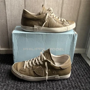 Philippe Model beige sneakers i skinn - Snygga beige sneakers från Philippe Model med klassisk låg profil och rund tå. Skorna är tillverkade i mjukt skinn med vita snören och vit platt sula. På sidan finns en broderad logotyppatch. Perfekt för dig som gillar stilrena och exklusiva sneakers.