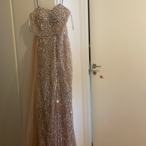 Champagnefärgad långklänning med paljetter - Säljer en magisk långklänning i champagnefärg med glittriga paljetter och snygg snörning i ryggen. Klänningen har tunna axelband och ett lager av mjuk tyll på sidan som ger extra volym. Perfekt för bal eller fest, du kommer verkligen sticka ut!