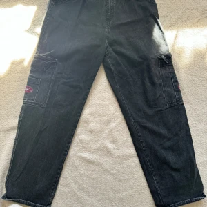 Svarta cargopants från Wake Jeans - Säljer ett par svarta cargopants från Wake Jeans med flera stora fickor på sidorna och bak, samt broderade loggor i rött och blått. Byxorna har raka ben och är tillverkade i jeansmaterial. Perfekta för dig som gillar streetwear och praktiska detaljer.