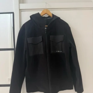 Svart tech-hood från Sail Racing - Svart tech-hoodie från Sail Racing med huva och två stora bröstfickor med lock. Jackan har dragkedja framtill och är tillverkad i 100% polyester. Snygg och sportig look med diskret logga på ena fickan. Perfekt för dig som gillar stilren och funktionell design.