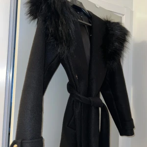 Svart kappa - Snygg kappa från River Island med bälte och konstpäls på axlarna. Perfekt för vintern och väldigt varm. Har bara använts en vintersäsong och finns inga skador på den. 