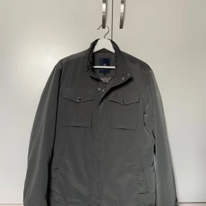 Grå/Grön vindjacka från Riley, strl 48 - Snygg grå/grönvindjacka från Riley med ståkrage och praktiska fickor framtill. Jackan har en stilren look med knappar på bröstfickorna och är tillverkad i ett slätt syntetmaterial. Perfekt för dig som gillar enkel och clean stil. Det finns några fläckar på ryggen men dom är inte särskilt synliga