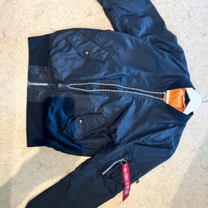 Svart bomberjacka från Alpha Industries - Säljer en klassisk svart bomberjacka från Alpha Industries med orange foder och ikonisk 'Remove Before Flight'-tag på ärmen. Jackan har dragkedja framtill, fickor med tryckknappar och ribbade muddar. Perfekt för dig som vill ha en streetig och tidlös look.