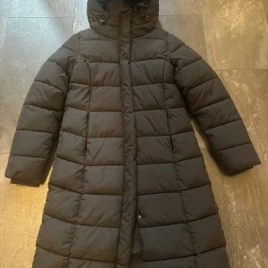 Svart lång pufferjacka från 157 XS - Säljer en svart lång pufferjacka från 157 i storlek XS. Jackan har huva med dragsko, dragkedja och tryckknappar framtill. Quiltad design med raka sömmar och lång passform som håller dig varm under kalla dagar.