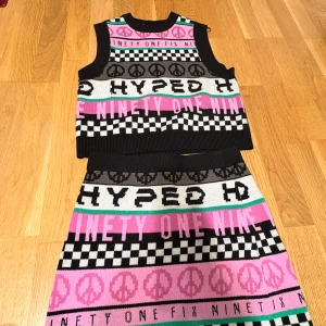 Färgglad stickad kjol med mönster - Säljer en cool stickad kjol och matchande tröja  med grafiska mönster, checkerboard-rutor, peace-symboler och text i svart, rosa, grått och vitt. Kjolen har en rak, kort passform och är perfekt för dig som gillar statement-plagg och streetstyle. Tröjan är i xs och kjolen i S 