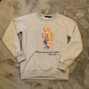 Grå sweatshirt Polo Bear Ralph Lauren - Grå sweatshirt från Ralph Lauren med ikoniska Polo Bear-trycket framtill. Tröjan har rund hals, långa ärmar och ribbade muddar. Tillverkad i mjuk bomullsmix, perfekt för en chill och stilren look.