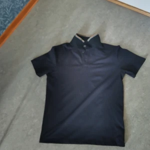 Mörkblå pikétröja med randig krage - Snygg mörkblå pikétröja från H&M med korta ärmar och klassisk krage. Kragen har en stilren randig detalj i beige och vitt. Tröjan har knappar vid halsen och är gjord i mjuk bomull som känns skön mot huden. Perfekt för en chill och stilren look.