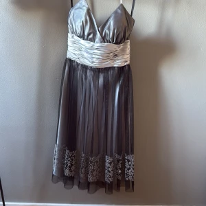 Silvergrå kort klänning från Morgan & Co - Snygg silvergrå klänning med omlott-v-ringad överdel och bred, glansig midja i ljus satin. Kjolen är kort med ett lager svart tyll och broderade blommor längst ner. Axelbandslös design och dragkedja bak. Perfekt för dig som vill sticka ut med elegans.